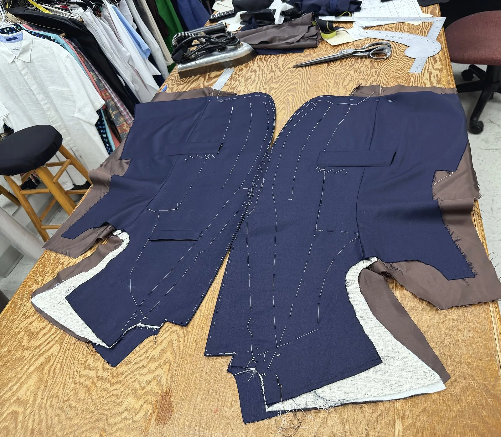 The workroom bench: basted navy jacket fronts, an iron, shears and a curve ruler, shirts on the rack behind
